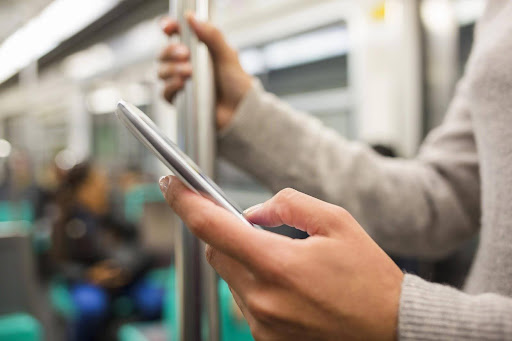 A close-up of an individual in business casual attire holding a smartphone in one hand while hanging onto a support post in a bus or subway car with the other; FLSA travel time compensation rules.
