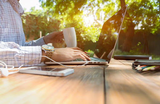 A man in a button-down shirt holds a cup of coffee while tapping on the keyboard of a laptop resting on a picnic table beneath a shade tree; telecommuters enjoy the protections of wage and hour laws.