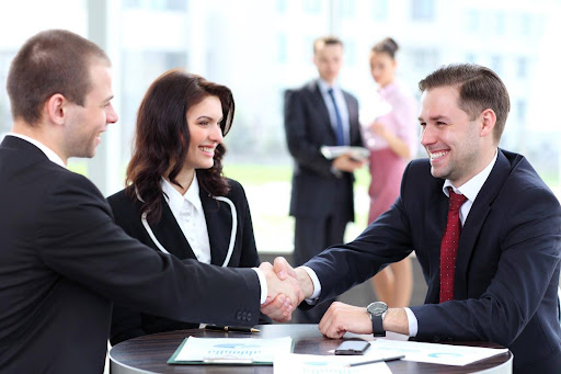 A transactional lawyer smiling as she watches two businessmen in suits shake hands over a table covered in spreadsheets; benefits of transactional law services.