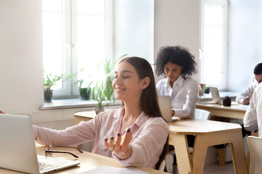 A woman participating in an employee wellbeing program focused on employee health. She is relaxed and content while her colleagues in the background focus on work. 