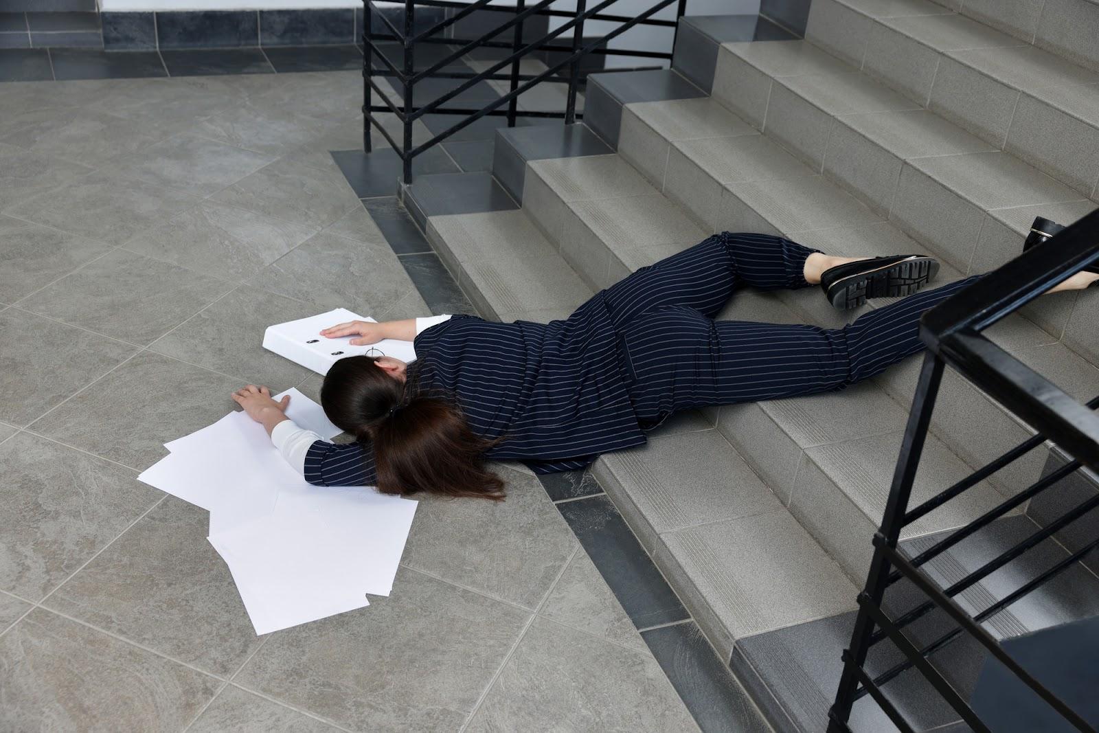 Woman in a pinstripe business suit and loafers lying face down unconscious across the bottom of a set of stairs, surrounded by scattered papers; premises liability concept. 