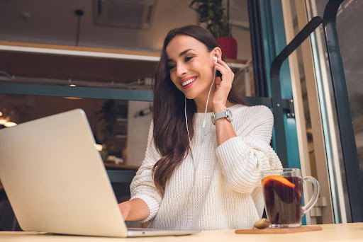 Young female employee seated at desk with a laptop and cup of tea, adjusting her earpiece as she takes a colleague’s call; a distributed workforce can significantly increase recruiting opportunities, but may add complexity to employers’ pay practices.