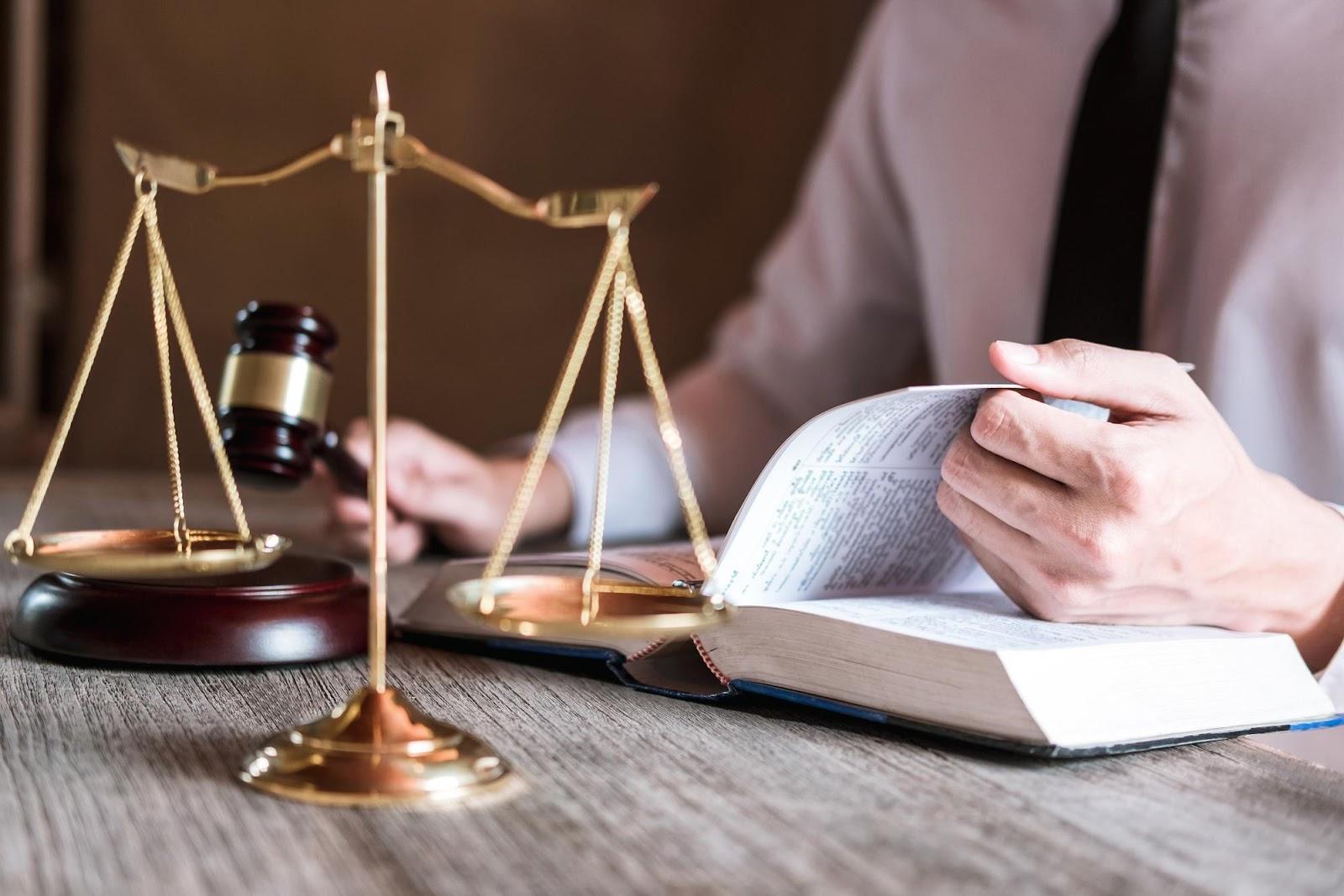 Closeup of a desk with the scales of justice; a man in business attire is flipping pages in a legal text on the desk with his left hand, has gavel raised to tap in his right.