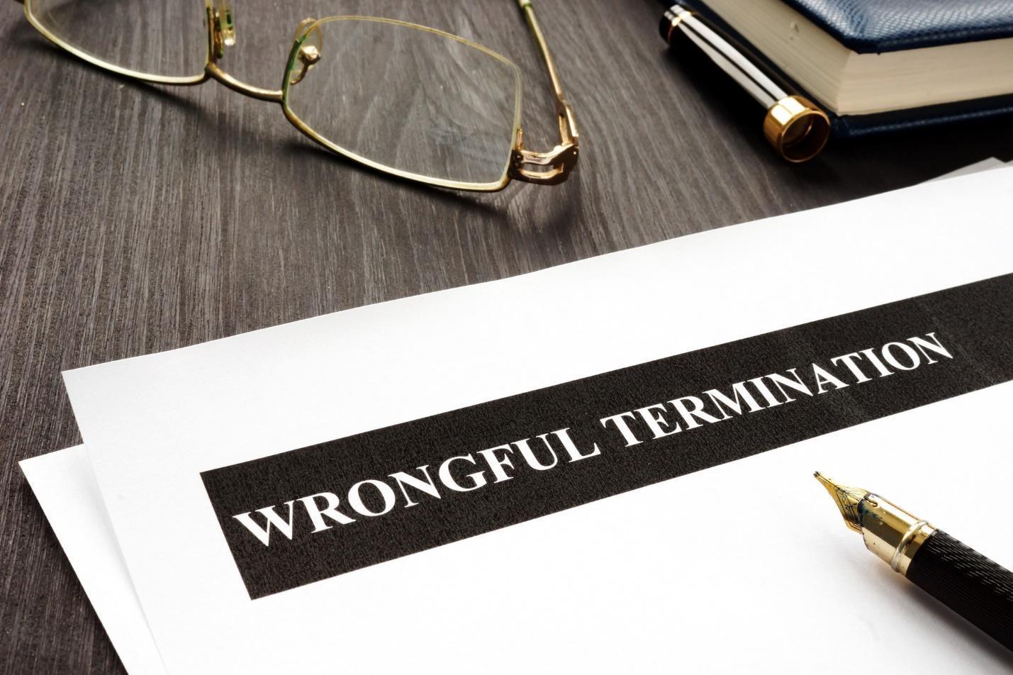 The top of a piece of paper with the words wrongful termination typed at the top. A pen sits on top of the paper, and a pair of glass is on the dark wood table above the paper.