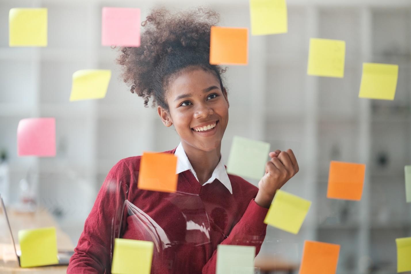 A smiling employee sits in front of a window with brightly-colored sticky notes on it as she improves her productivity under employee productivity monitoring.