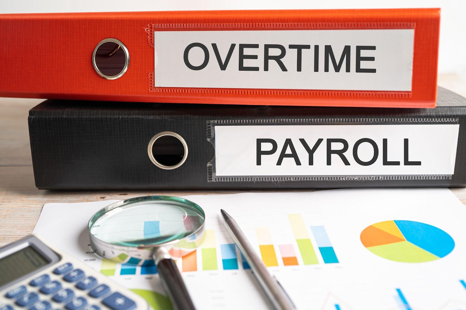 A red binder labeled “Overtime” sits on top of a black binder labeled “Payroll,” next to a calculator, magnifying glass, pen, and page filled with charts. Avoid wage and hour class action lawsuits with simple risk management strategies such as keeping accurate track of employees’ working hours.